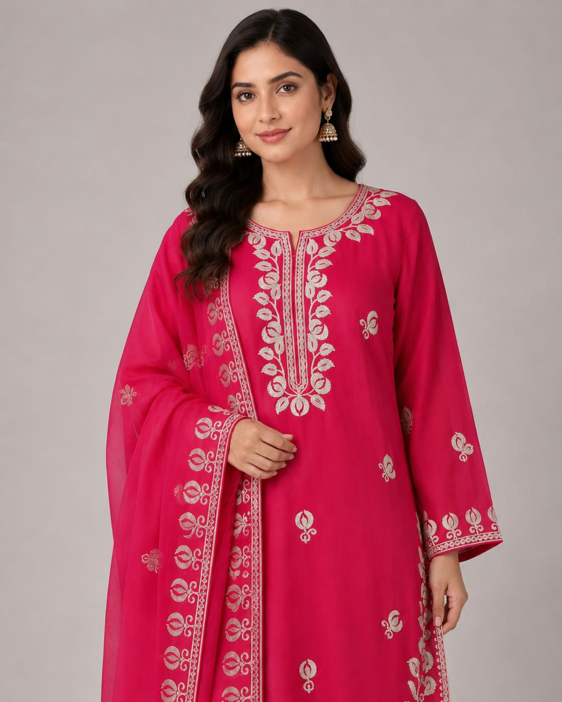 A woman modeling a bright pink traditional Indian outfit with a matching sheer dupatta, featuring intricate white floral embroidery on the kurta and dupatta, paired with matching pink trousers, against a neutral gray studio background.
