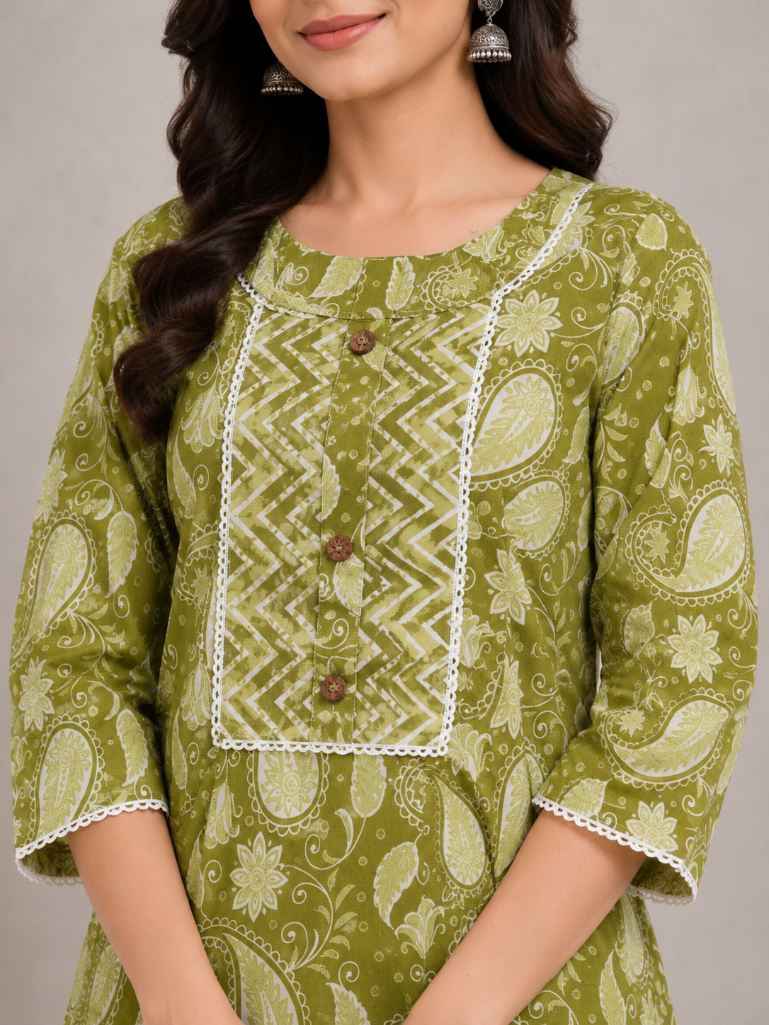 A woman models a green printed kurta set with matching patterned pants, standing in a studio setting with a neutral gray background and a small plant on a white pedestal.