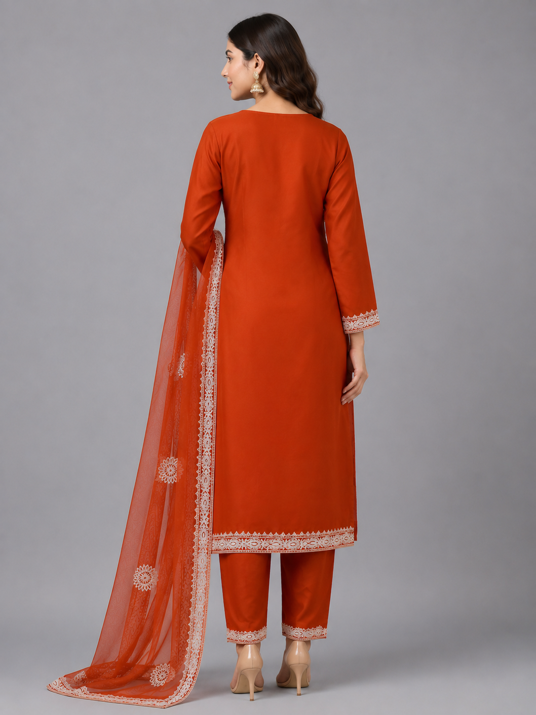 A woman modeling a rust-orange traditional Indian salwar kameez suit with white embroidery and a matching sheer dupatta, set against a solid gray studio background.