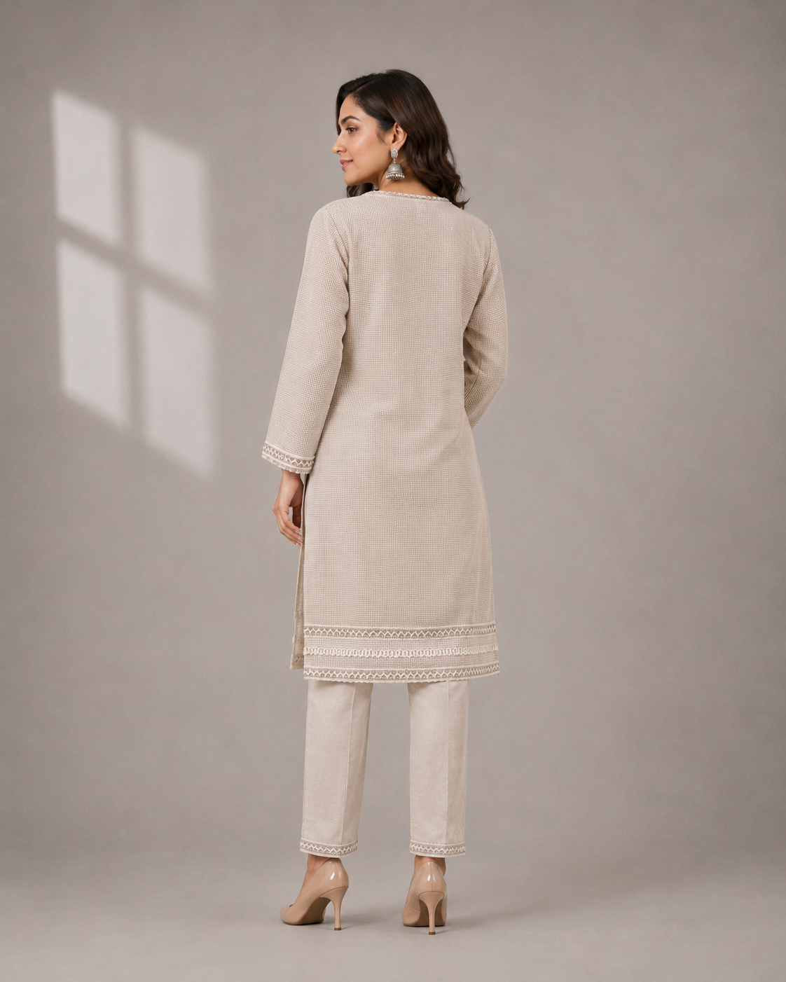 A woman modeling a light beige kurta set with matching trousers, featuring delicate gold embroidery on the neckline, cuffs, and hem, set against a solid warm beige studio background.