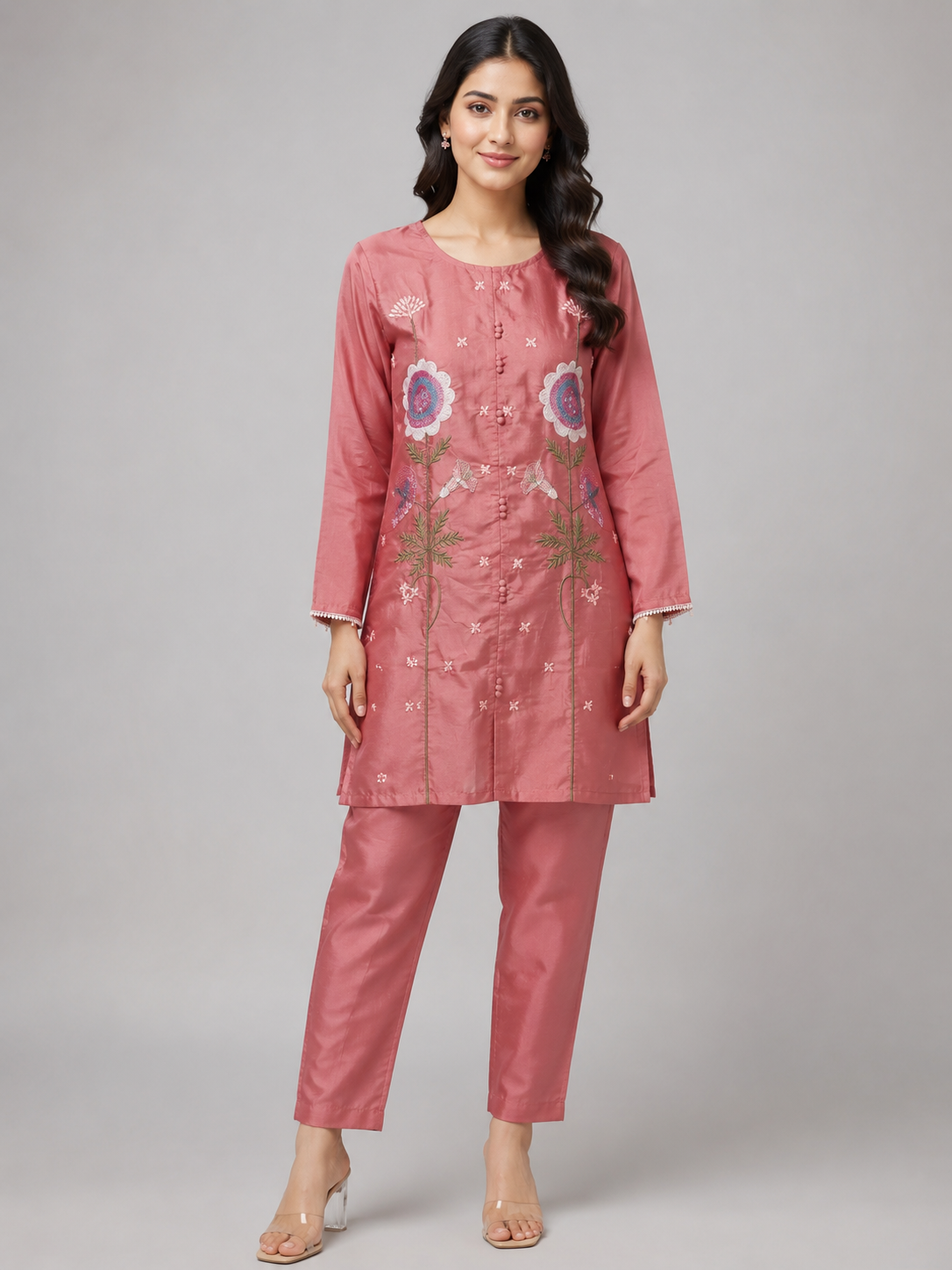 A woman modeling a dusty pink kurta and trouser set with floral embroidery on the front, photographed against a neutral gray studio background.
