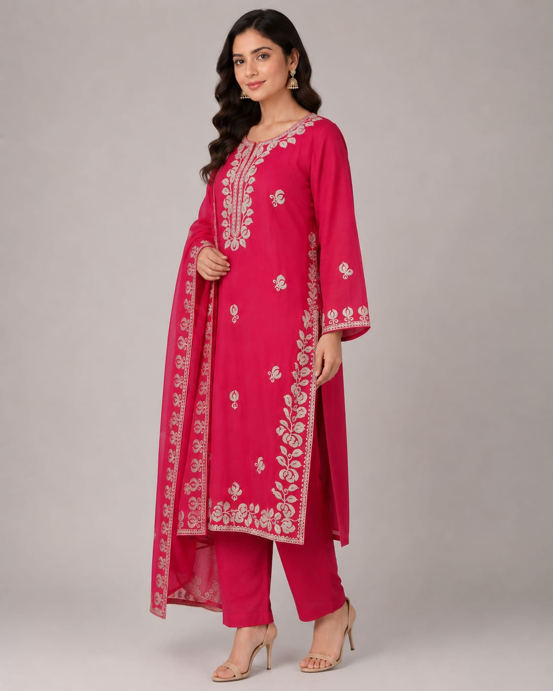 A woman modeling a bright pink traditional Indian outfit with a matching sheer dupatta, featuring intricate white floral embroidery on the kurta and dupatta, paired with matching pink trousers, against a neutral gray studio background.
