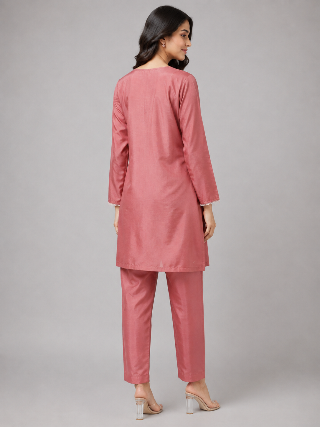 A woman modeling a dusty pink kurta and trouser set with floral embroidery on the front, photographed against a neutral gray studio background.