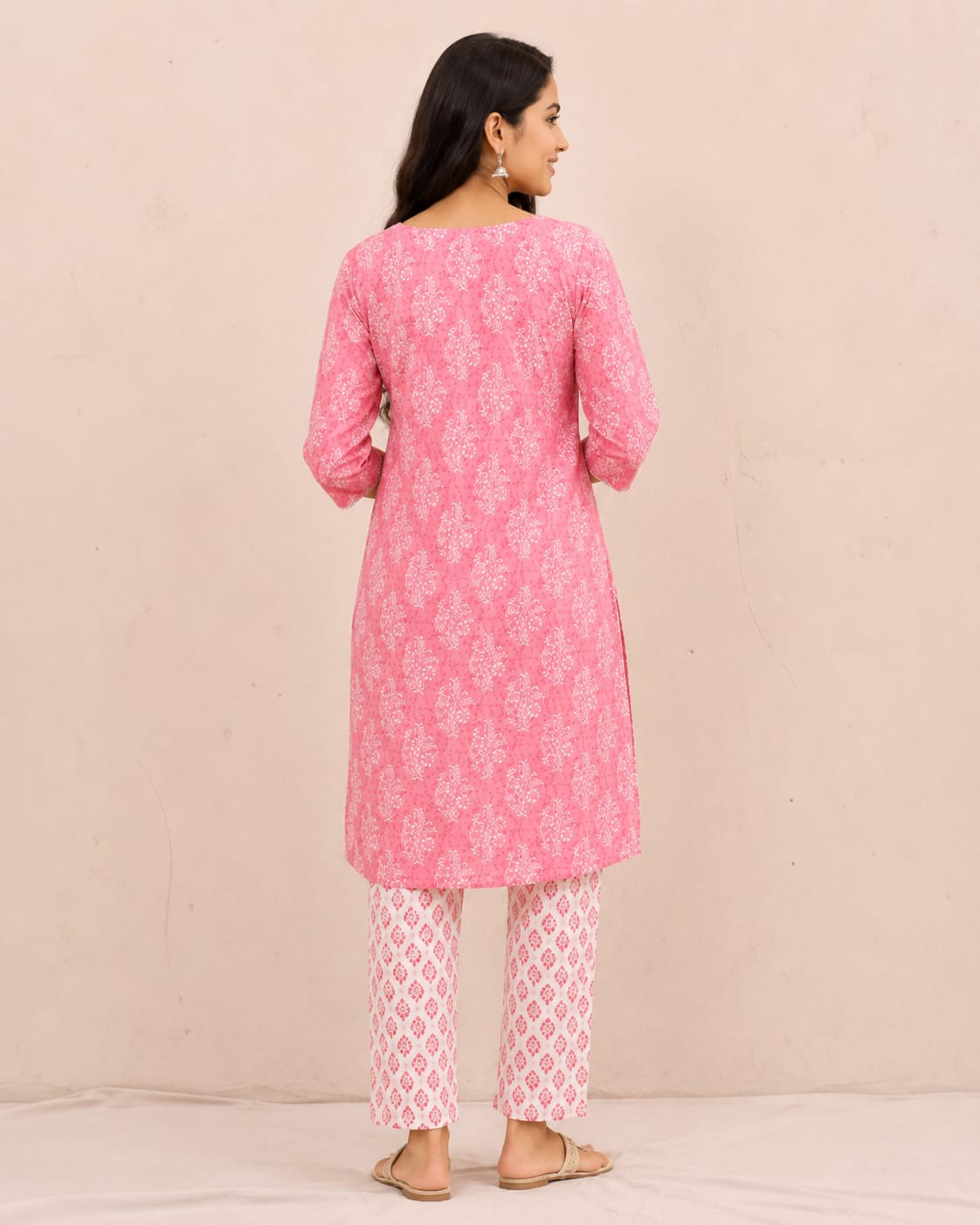 A woman modeling a pink printed kurta with white embroidered detailing on the chest, paired with matching pink and white patterned pants, against a solid light beige background.