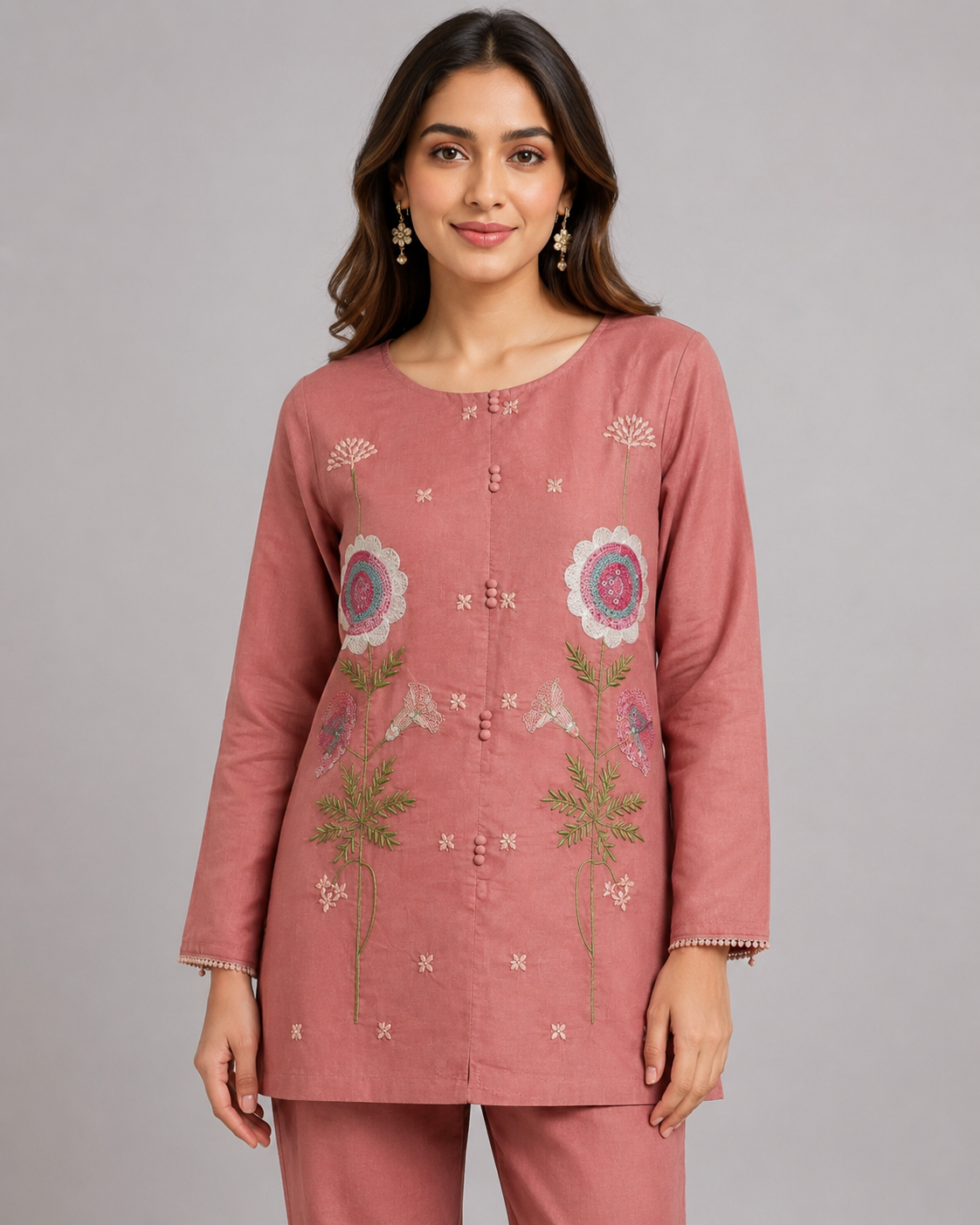 A woman modeling a dusty pink kurta and trouser set with floral embroidery on the front, photographed against a neutral gray studio background.