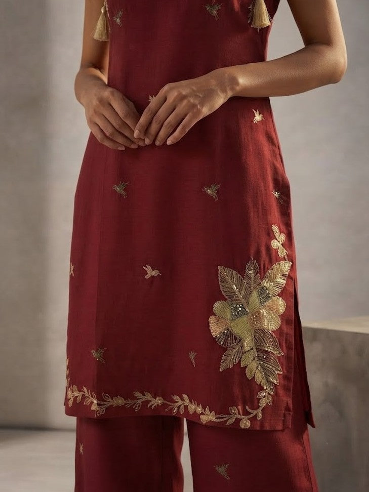 A woman models a deep red kurta set with wide-leg pants, featuring gold floral embroidery on the neckline, hem, and kurta front, set against a minimalist concrete block studio background.