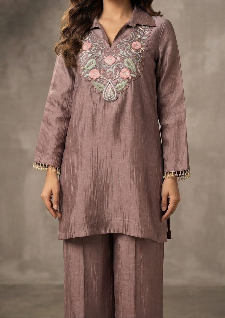 A woman modeling a dusty mauve two-piece kurta and trouser set with floral embroidery on the chest, standing against a textured gray studio backdrop.