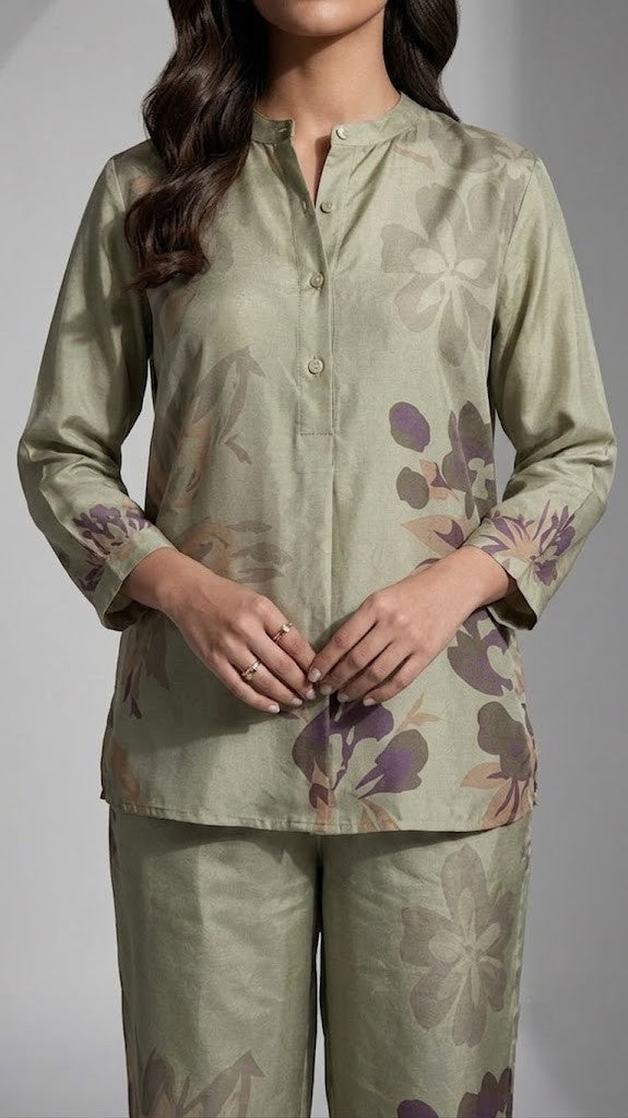 A woman modeling a two-piece satin pajama set in sage green with a floral print, posed against a gray studio background with a circular spotlight effect.